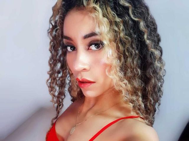 KylieJen has curly blonde and brown hair, red lipstick, and a red top with a gold necklace, looking at the camera with a slight smile.