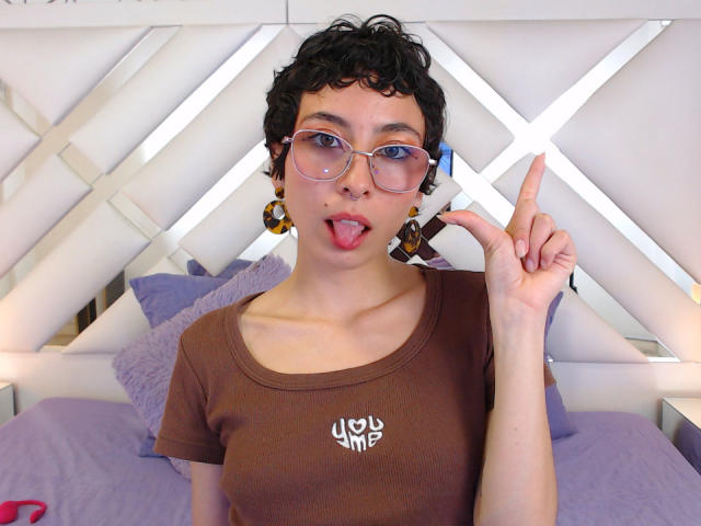 EmilyAllen with curly black hair, glasses, and brown t-shirt, sticking out tongue, pointing up with finger, small nose ring, and colorful earrings, looking cute and playful.