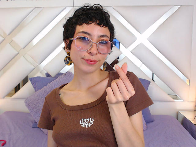 EmilyAllen with curly black hair, blue glasses, brown t-shirt, and gold earrings, pointing with her index finger, smiling, on a bed with purple pillows.
