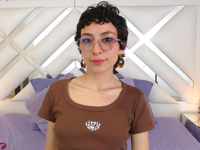 EmilyAllen has short curly black hair, wears purple glasses, and a brown t-shirt with white text. She's smiling with purple lips and orange earrings, showing her small tits.