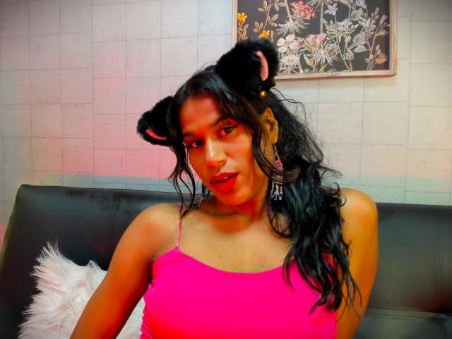 GodessMar with black bear ears, pink tank top, and long wavy hair, sitting on black leather couch, pink light, fluffy pillow, and floral wall art in background.