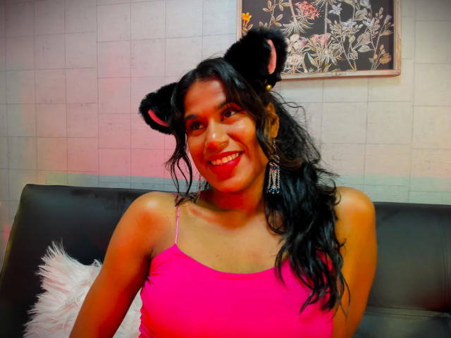GodessMar with black furry bear ears, pink tank top, long wavy black hair, smiling, red lipstick, white fluffy pillow, black leather couch, and floral wall art.
