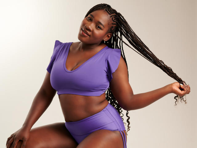 MissBNastyX, avec sa peau foncée, ses longs cheveux tressés, son haut court violet et sa culotte haute assortie, tenant ses cheveux, souriant, montrant ses seins et ses fesses, debout devant un fond uni..