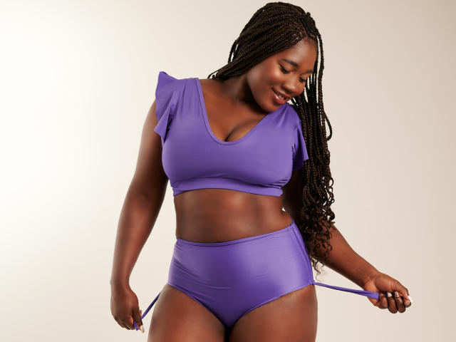 A photograph of MissBNastyX with dark brown skin and long, braided hair. She wears a purple two-piece outfit with a short-sleeved crop top and high-waisted bottoms. She holds the waistband of her bottoms with her right hand, slightly pulling it. Her left arm is relaxed by her side. She looks down with a slight smile. The purple outfit contrasts with her dark skin. The image has a clean, minimalist background. Her midsection is toned, and she has a confident, relaxed posture.