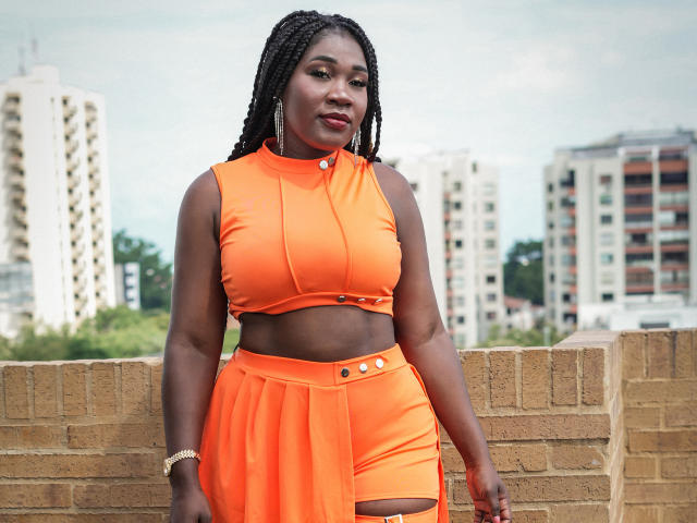 MissBNastyX met donkere huid, gevlochten haar, een oranje topje en hoge shorts, waarbij haar billen en kleine borsten zichtbaar zijn, staat tegen een bakstenen muur, en draagt een gouden armband..