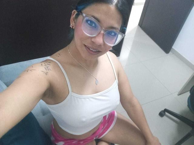 Paulina Colemann, vêtue d'un débardeur blanc, de shorts roses et portant des lunettes, a les cheveux bleus, de petites seins et un tatouage sur son épaule droite, et elle est assise sur une chaise..
