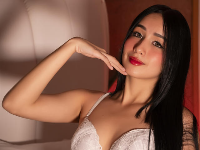 Valentina Risso, con i suoi lunghi capelli neri, il rossetto rosso e il reggiseno di pizzo bianco, che si tocca il mento con la mano destra, sorridendo leggermente, mostrando il seno e i glutei..