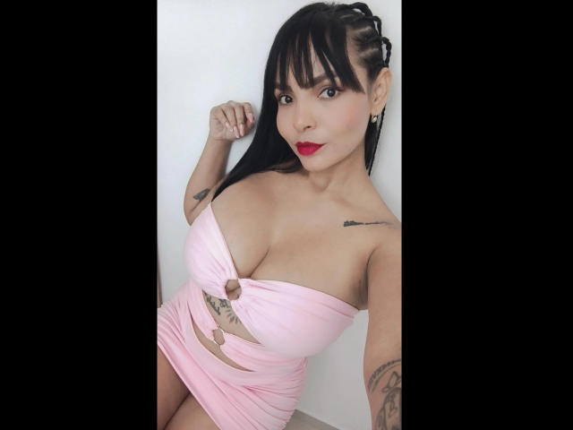Jessica Fiore, avec de grandes seins, portant une robe rose avec une découpe, a des tatouages sur les bras, du rouge à lèvres, et des cheveux noirs avec une frange, et elle se tient devant un mur blanc..