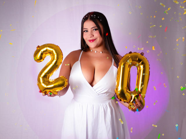 LorensDouce with big tits, wearing a white dress, holding gold "20" balloons, smiling, confetti around, pink and purple light, dark hair, red lipstick, white necklace.