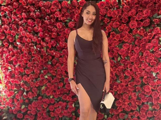 AmelieClair stands in a black dress with a thigh-high slit, holding a white purse, surrounded by a wall of red roses, wearing a black wristwatch, smiling.