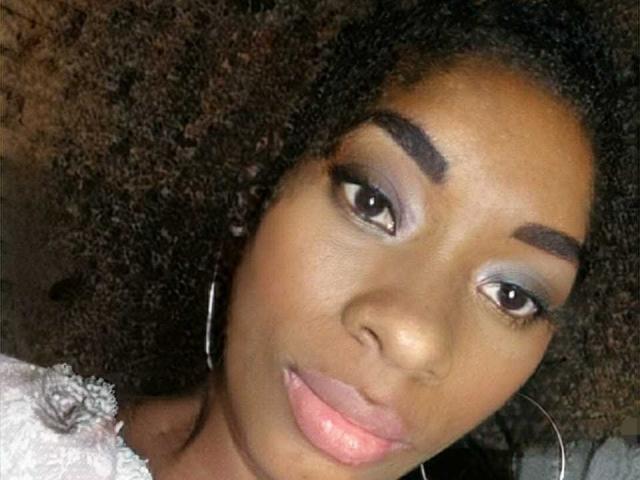 Shaaron's face with dark brown skin, curly black hair, bold eyebrows, blue eyeshadow, long eyelashes, pink lipstick, and silver hoop earrings, looking slightly to the side.