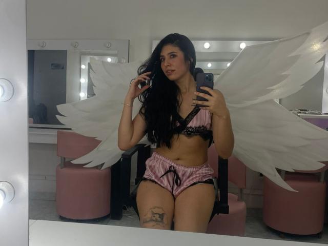 MiluSky with black hair, pink and black lace bra, pink satin shorts, white angel wings, holding phone, mirror selfie, tattoo on right thigh, pink chair, bathroom lights.