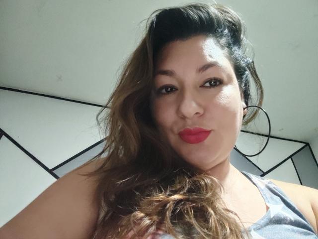 SakotoDika mit welligem, braunem Haar, rotem Lippenstift, Reifenohrringen und einem grauen Tanktop, die in die Kamera lächelt. Ihre Brüste sind teilweise sichtbar..