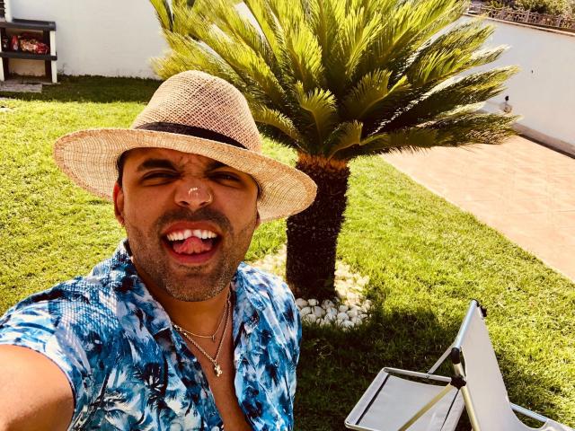 TrioTemptation stands in a sunny backyard, wearing a blue tie-dye shirt and a beige straw hat. He has a medium build, a beard, and is smiling with his tongue out. His eyes are squinted, and he wears a silver necklace with a small pendant. A palm tree is behind him, and a white chair is in the foreground. The green grass and white wall are visible. Bright sunlight creates strong shadows. The image has a casual, summery vibe.