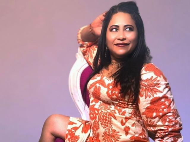 MiaSalomeChaude with dark hair, wearing an orange floral dress, sitting on a white chair, looking to the side, hand in hair, smiling, visible tits, light brown skin.