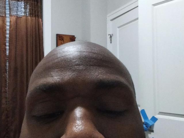 Shauncampbell-hot's bald head with shiny, dark brown skin, closed eyes, and slightly raised eyebrows, showing a smooth, shiny texture. A white door and brown curtain are in the background.