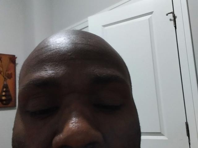 Shauncampbell-hot's bald, dark-skinned face with closed eyes, visible eyebrows, and slight smile. White door in background, left-side wall art with vase.