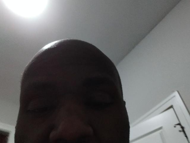 Shauncampbell-hot's bald head and dark eyes are visible in a low-light, dimly lit room with a ceiling light and a white door in the background.