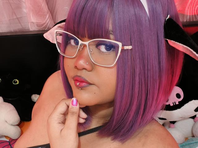 Close-up of MissCurvyTop with purple and pink hair, wearing white-framed glasses, and black cat ears. She has a light brown skin tone, a small nose piercing, and pink lipstick. Her right hand, with purple nail polish, touches her chin. She's wearing a black strap. The background includes stuffed animals. The image has a soft, colorful, and playful style. The focus is on her face and glasses, with a slightly tilted head. Her expression is neutral.
