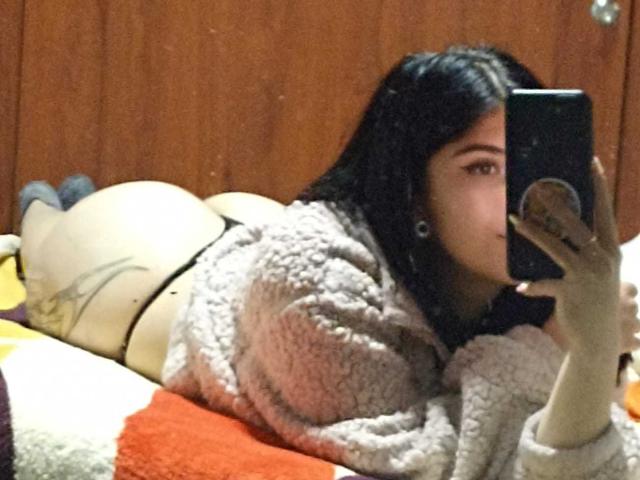 EmmaCollinsHot lies on a bed, wearing a fluffy beige robe, black thong, and holding a phone, showing her bare ass, with long dark hair and a neutral expression.