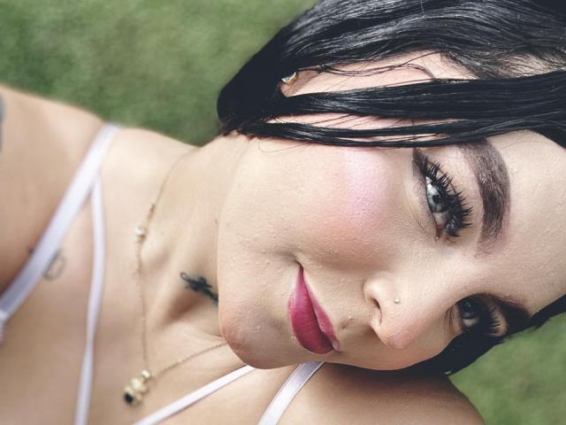 Il volto di MelodyBlossom è presente nella foto, con capelli neri, occhi verdi, rossetto rosa e un leggero sorriso. Indossa una bretella bianca, e si può notare un piccolo tatuaggio..