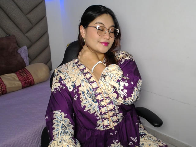 HelueJamil, Aziatische vrouw met een bril, paars en wit geborduurde jurk, parelketting, zwart haar, lachend, zittend in een zwarte stoel, handen in schoot, bed met kussen op de achtergrond..