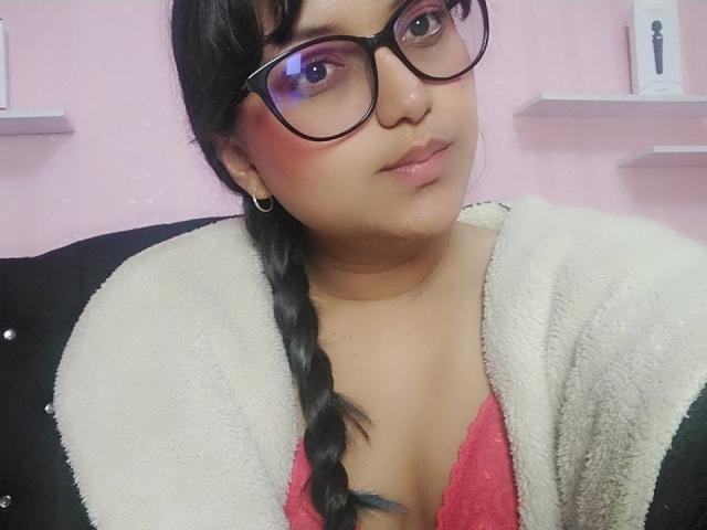 AzulMoon, com tranças escuras, óculos e blush rosa, vestindo um cardigã branco felpudo sobre um sutiã de renda vermelho, mostrando o decote, um brinco de argola preto e um leve sorriso..