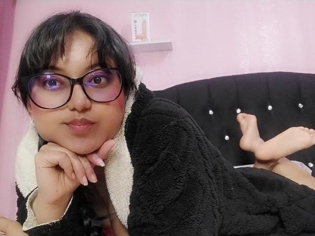 AzulMoon, avec ses cheveux noirs, ses lunettes violettes et sa robe noire et duveteuse, est allongée sur un lit noir capitonné, la tête reposant sur ses mains, affichant un sourire mignon et les pieds nus..