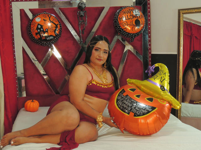 MicaelaHot, chubby with big ass, wearing a red bikini, gold jewelry, and a headpiece, sits on a bed with a Halloween pumpkin balloon and a witch hat.