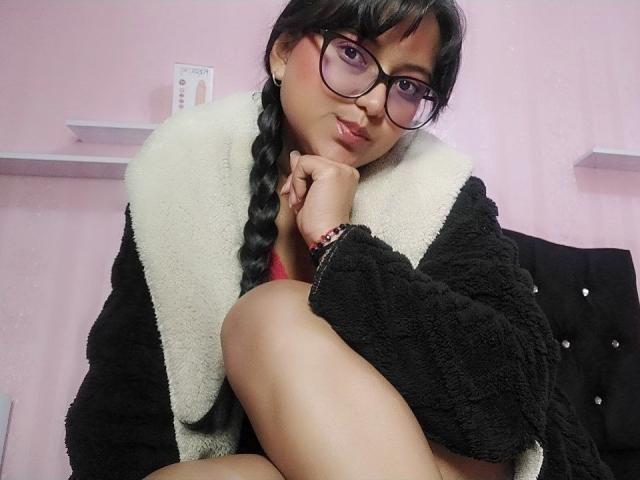 AzulMoon, com cabelo trançado escuro, óculos e batom rosa, vestindo um casaco preto e branco felpudo, sentada com os joelhos dobrados, olhando para a câmera com um sorriso discreto..