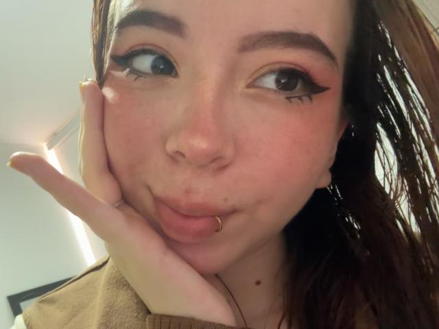 A KattyHards tem um rosto bonito, com olhos castanhos, sombra rosa e um piercing no nariz. Ela está fazendo uma expressão de lábios franzidos, com a mão apoiada na bochecha. Seus lábios estão ligeiramente curvados para cima..
