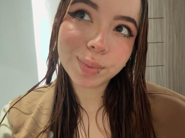 KattyHards, avec ses cheveux bruns mouillés, son rouge à lèvres rose discret, son eye-liner noir, son petit anneau de nez et un léger froncement de lèvres, portant un haut marron et regardant vers le haut..