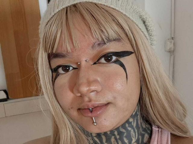 Close-up of DarkCurvyLady with light blonde hair, wearing a knitted gray beanie. She has dark, thick, black eyeliner extending into a winged pattern on her upper eyelids. Her face has three small metal piercings on her forehead above her nose. She has a silver lip piercing on her bottom lip. Her tattooed neck is visible, showing a detailed, dark, symmetrical pattern. She has a light skin tone with visible freckles and a small mole on her right cheek. Her expression is neutral. The background is minimal and indoor.
