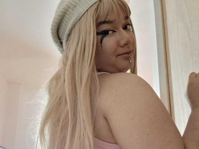 Donkere, rondachtige vrouw met lang blond haar, die een roze top draagt, een grijze muts, zwarte oogmake-up, een neusring en een lipring, en die over haar schouder kijkt. Haar billen zijn zichtbaar..