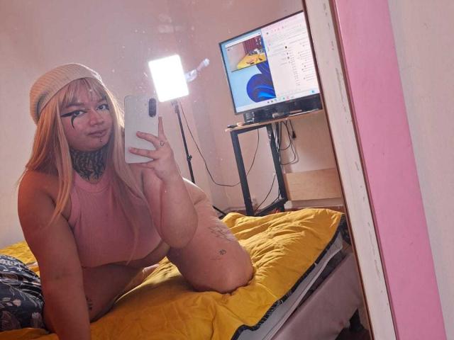 Donkere, rond gevormde vrouw met lang blond haar, een roze topje, een muts en tatoeages, knielend op een geel bed, die een spiegelzelfportret maakt met haar telefoon, waarbij haar blote borsten en billen te zien zijn..