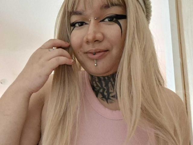 Una donna con lunghi capelli biondi, trucco occhi nero, piercing al naso, piercing al labbro inferiore e un tatuaggio sul collo, indossa una canotta rosa e sta toccando il viso..