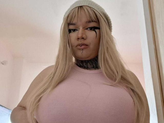 Donna dalla carnagione scura, con capelli biondi, berretto e eyeliner nero, che indossa una maglia rosa a coste, mostrando il suo seno prosperoso e un piercing al labbro, in una ripresa dal basso..