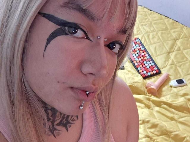 Close-up of DarkCurvyLady with blonde hair, black eye makeup, and a black eyebrow tattoo. She has a nose piercing, lip piercing, and a tattoo below her chin. Her pink tank top is visible. A yellow quilted bedspread, a grid of colorful tiles, and a pink vibrator are in the background. Her expression is neutral. The photo has a casual, personal feel. DarkCurvyLady's skin is fair, and her eyes are blue. The image is centered on her face.