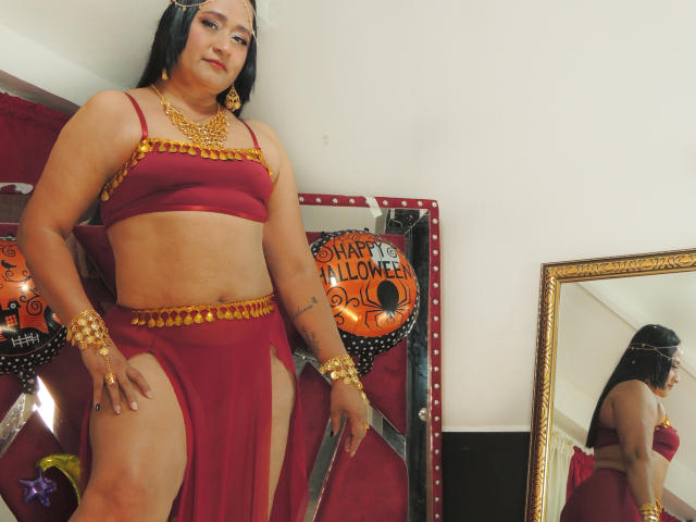 MicaelaHot with tan skin, black hair, red belly-dancer outfit, gold jewelry, showing her tits and pussy through slits, standing in front of a mirror, looking confident.