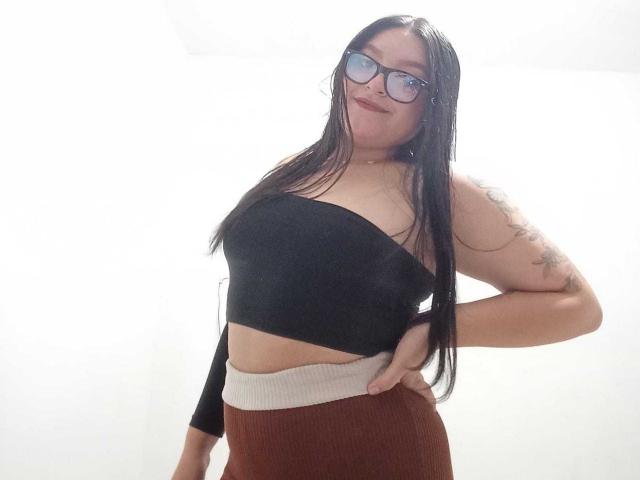 VictoriaRiver with long black hair, glasses, black strapless top, tan midriff, brown ribbed pants, tattoo on right arm, hand on hip, smiling, white background.