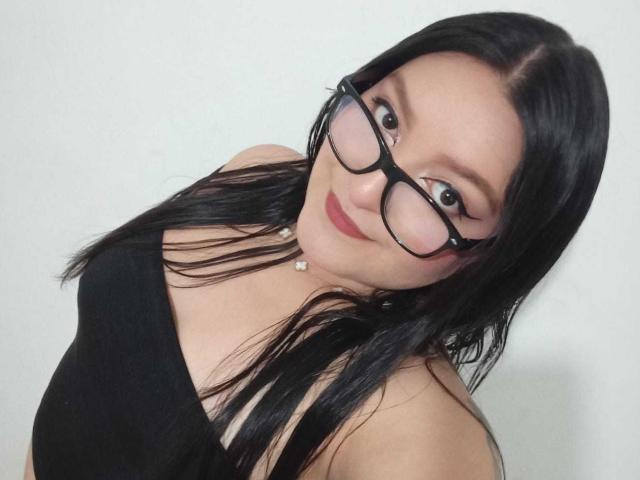 VictoriaRiver with black glasses, straight black hair, red lipstick, and a black tank top, showing a cute smile, looking up with a slight tilt. Boobs visible under tank top.