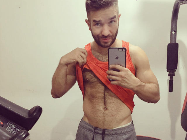 Niklaus, muscular, bearded guy with short brown hair, lifting his red tank top to show his hairy chest and abs, taking a mirror selfie with his iPhone.
