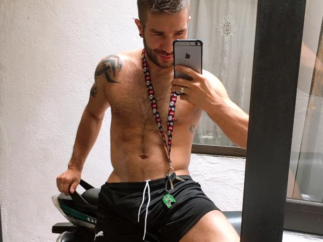 Niklaus, shirtless, muscular, hairy chest, black shorts, beaded necklace, tattoos on shoulder, holding phone in mirror, smiling, sitting on exercise bike, mirror reflection.