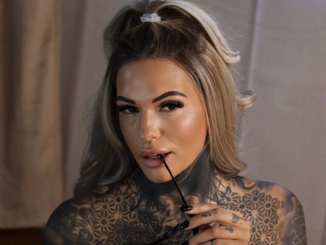 Aileenink-hot with tattoos, holding a black eyeliner with her teeth, has blonde hair in a ponytail, heavy makeup, and a nose ring, looking seductively at the camera.