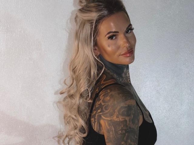 Aileenink-hot with long blonde curls, tattoos on her neck and arms, looking over her shoulder, wearing a black top, smiling with red lips.