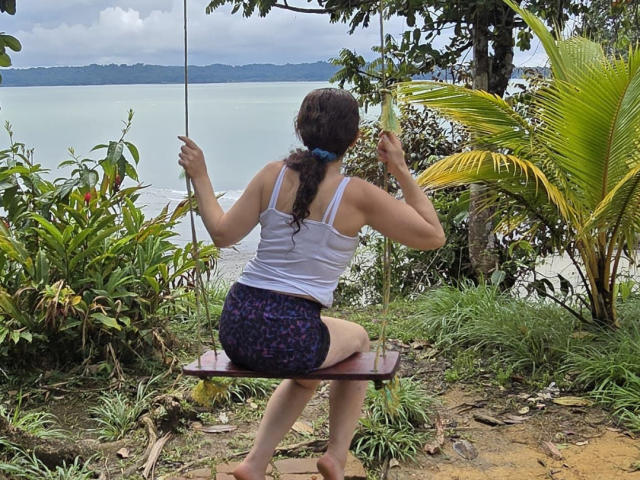 SamaraBellet, back to camera, sitting on a wooden swing, white tank top, purple leopard print shorts, brown hair in a braid, looking at a calm, greenish-blue lake.