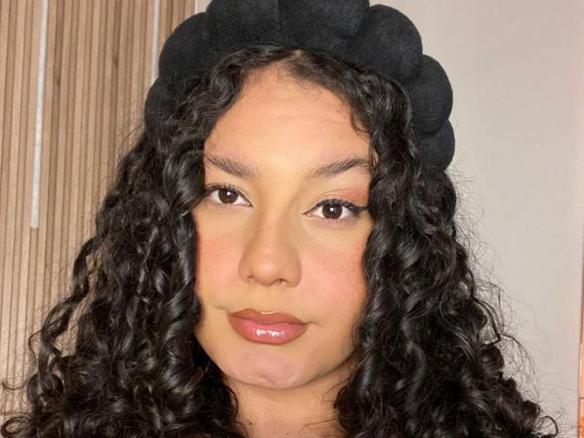 RachellBeaumont has curly black hair, a black fluffy hat, brown eyes, and glossy pink lips. Her light brown skin has a slight blush. Her tits are not visible.