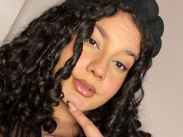 RachellBeaumont has curly black hair, a black beret, and glossy pink lips, with a finger gently touching her chin, brown eyes, and a soft, slightly smiling expression.