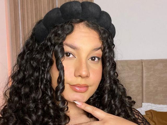 RachellBeaumont with curly black hair, black beret, brown eyes, and glossy pink lips, touching her chin, looking at the camera, in a bedroom with a beige headboard.