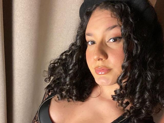 RachellBeaumont has curly black hair, wearing a black hat, with glossy pink lips, brown eyes, and a slight pout, showing her cleavage in a zip-up top.